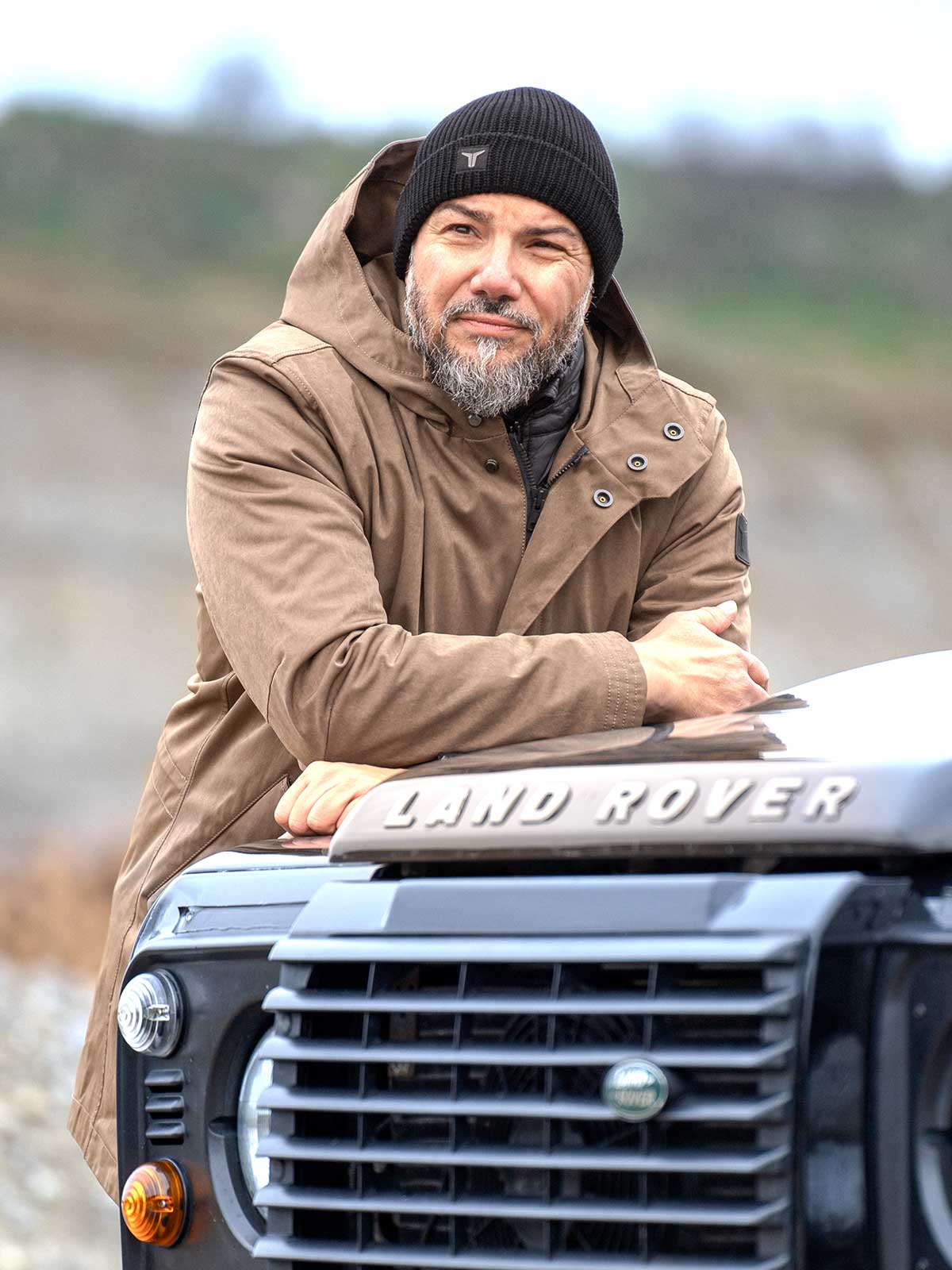 Model wearing a premium winter parka for men from for-legends.com by Caduff, crafted from exclusive high-density knit Japanese fabric, standing in a rocky winter landscape in the background a Land Rover Defender. / Modell trägt einen hochwertigen Winterparka für Herren von for-legends.com by Caduff aus einem exklusivem High-Density-Jersey aus Japan, stehend in einer felsigen Winterlandschaft im Hintergrund ein Landrover Defender.