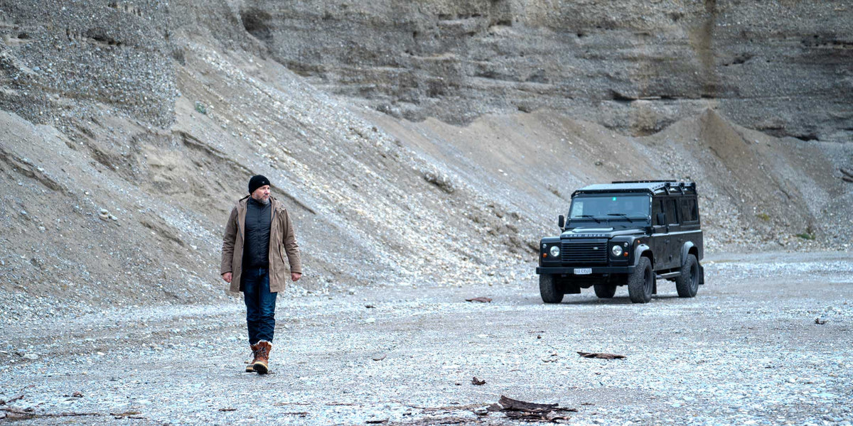 Model wearing a premium winter parka for men from for-legends.com by Caduff, crafted from exclusive high-density knit Japanese fabric, standing in a rocky winter landscape in the background a Land Rover Defender. / Modell trägt einen hochwertigen Winterparka für Herren von for-legends.com by Caduff aus einem exklusivem High-Density-Jersey aus Japan, stehend in einer felsigen Winterlandschaft im Hintergrund ein Landrover Defender.