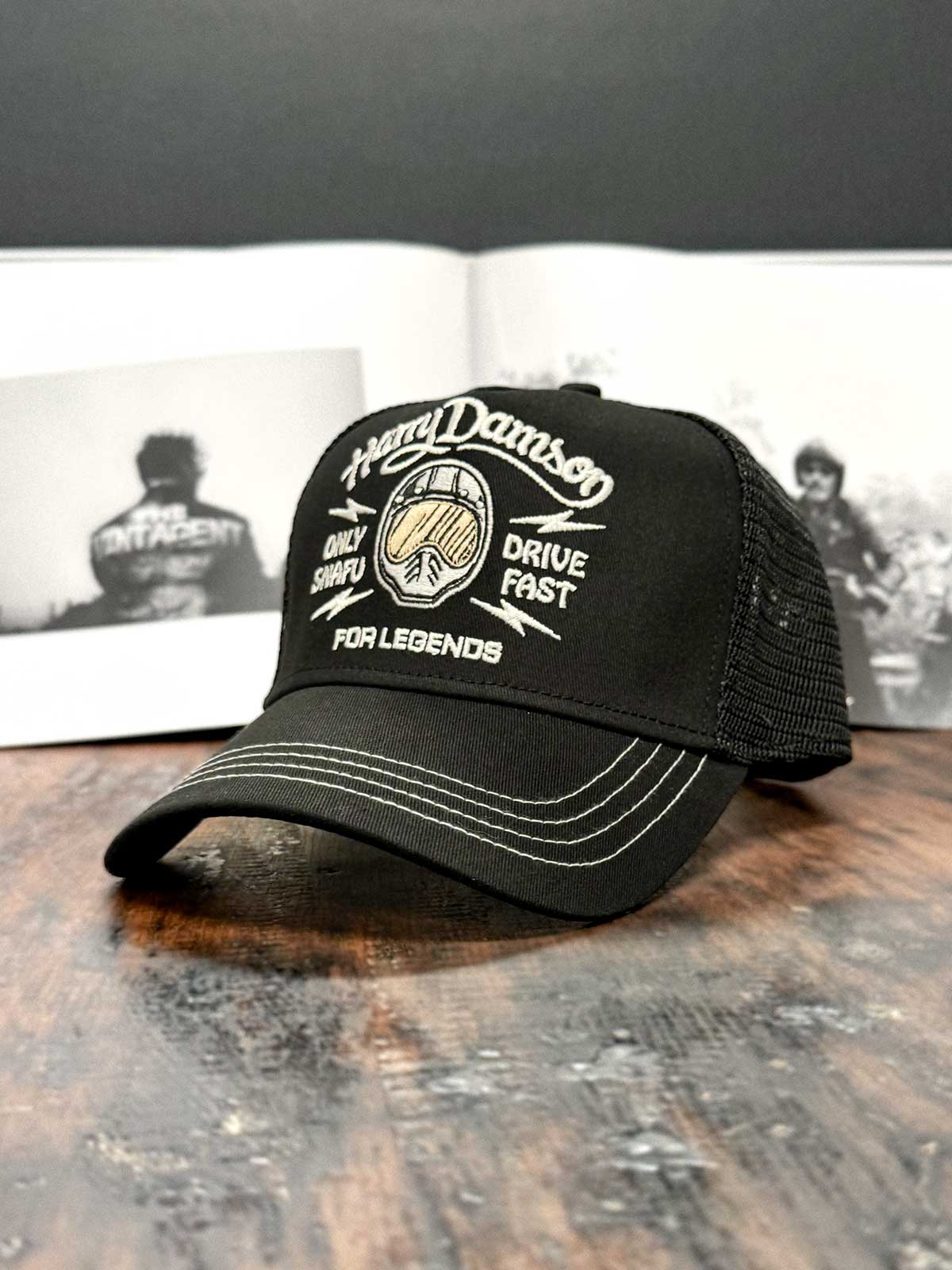 Schwarze Trucker Cap in schräger Seitenansicht von links, mit Motorradhelm und 'Only Snafu - Drive Fast' Schriftzug im Mittelpunkt, For Legends x Harry Damson