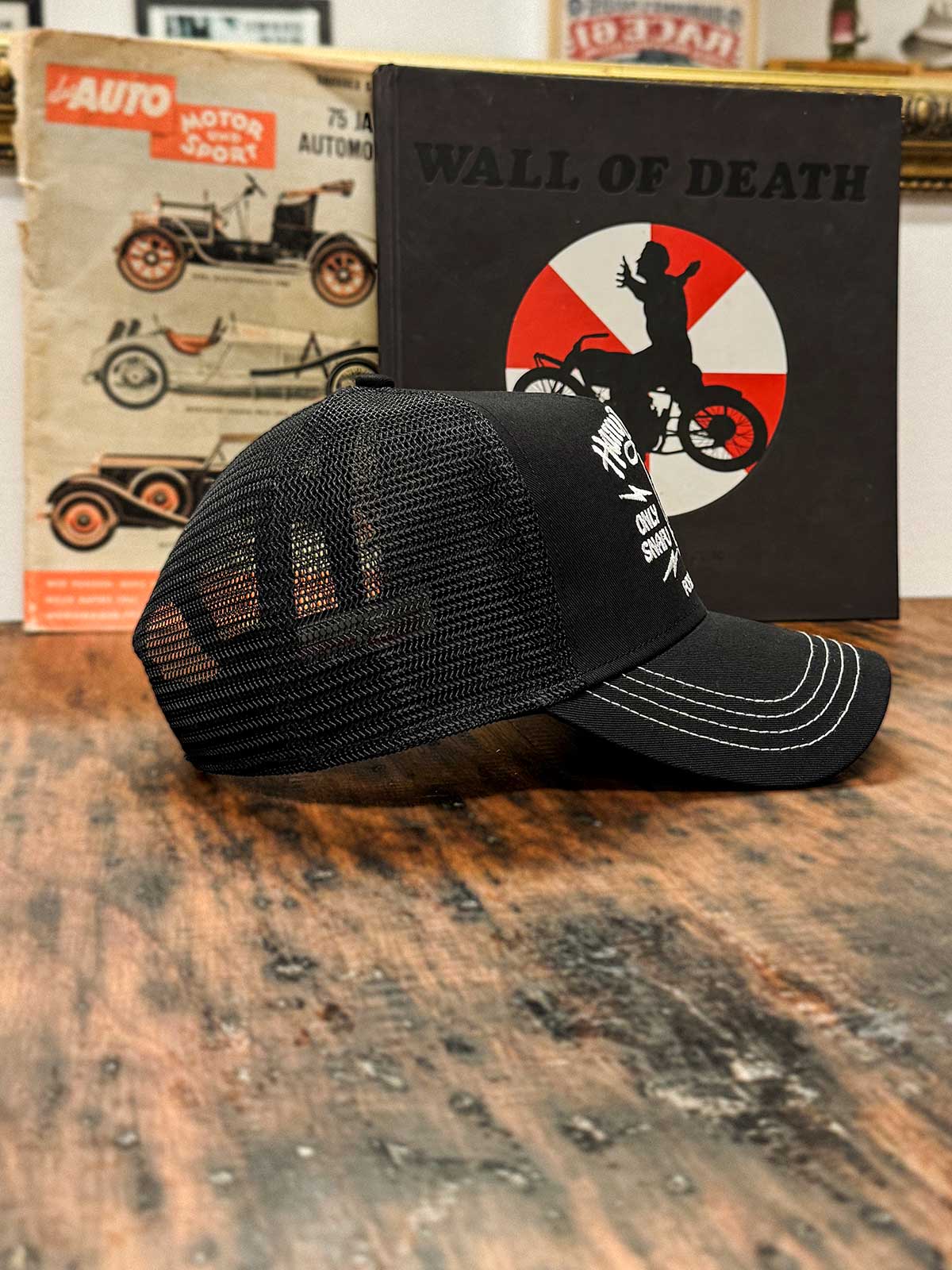 Schwarze Trucker Cap von der Seite, mit dem Buch 'Wall of Death' im Hintergrund, For Legends x Harry Damson