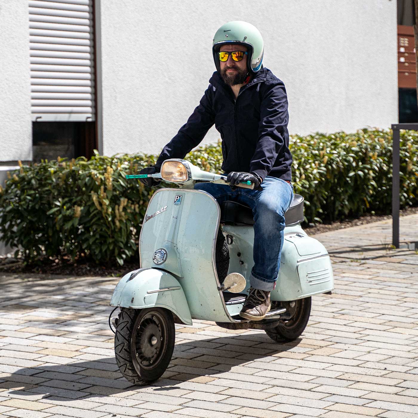 Ein Mann sitzt auf einer Vespa, lacht und trägt einen abriebfesten Parka der Marke For-Legends by Caduff, ideal für urban mobility - for-legends.com. A man sits on a Vespa, laughs and wearing an abrasion-resistant parka by For-Legends by Caduff, ideal for urban mobility - for-legends.com.