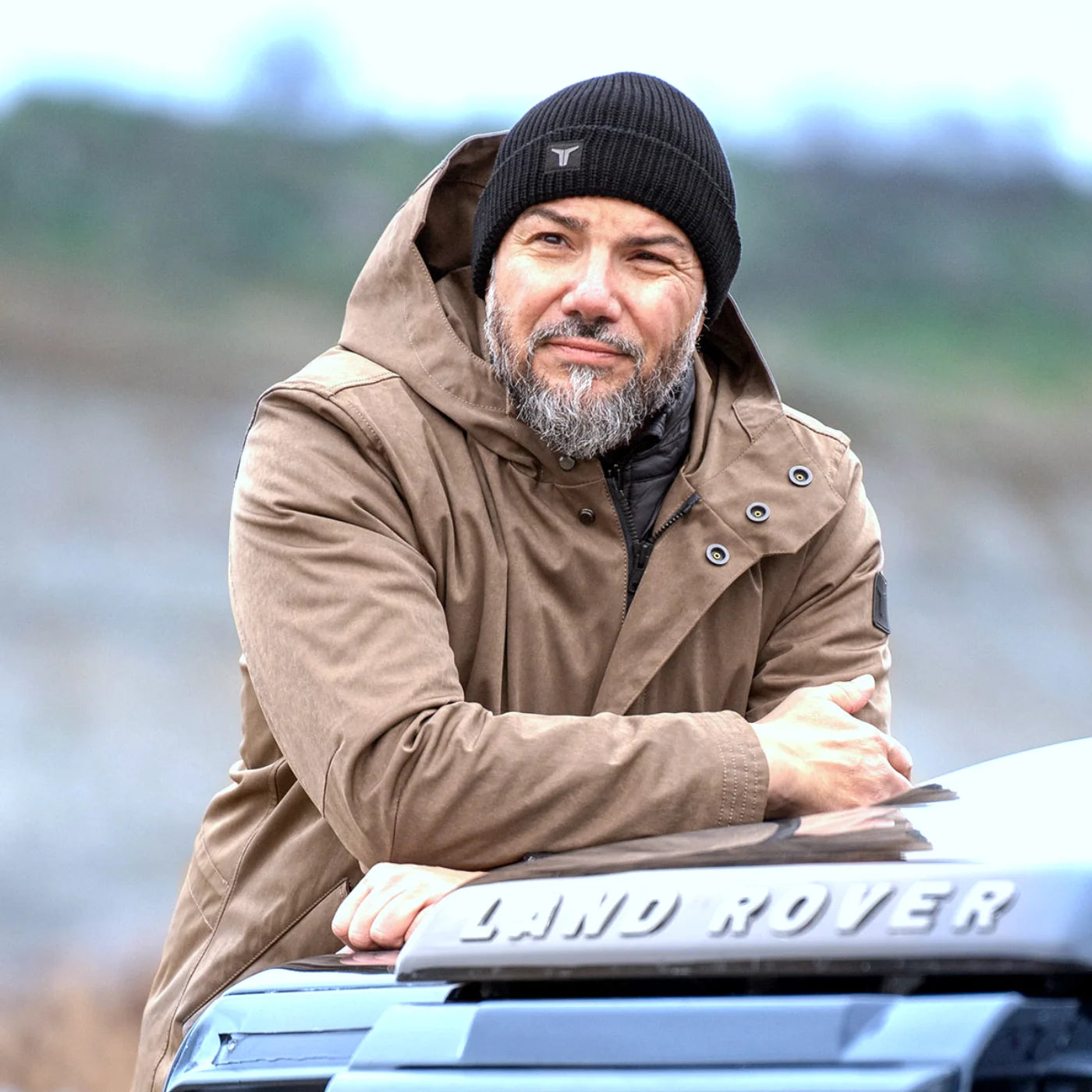 Model wearing the Brown CADUFF Japanese Double-Knit 2-in-1 Winter Parka leaning on a Land Rover Defender; YKK snap buttons visible on the front placket.