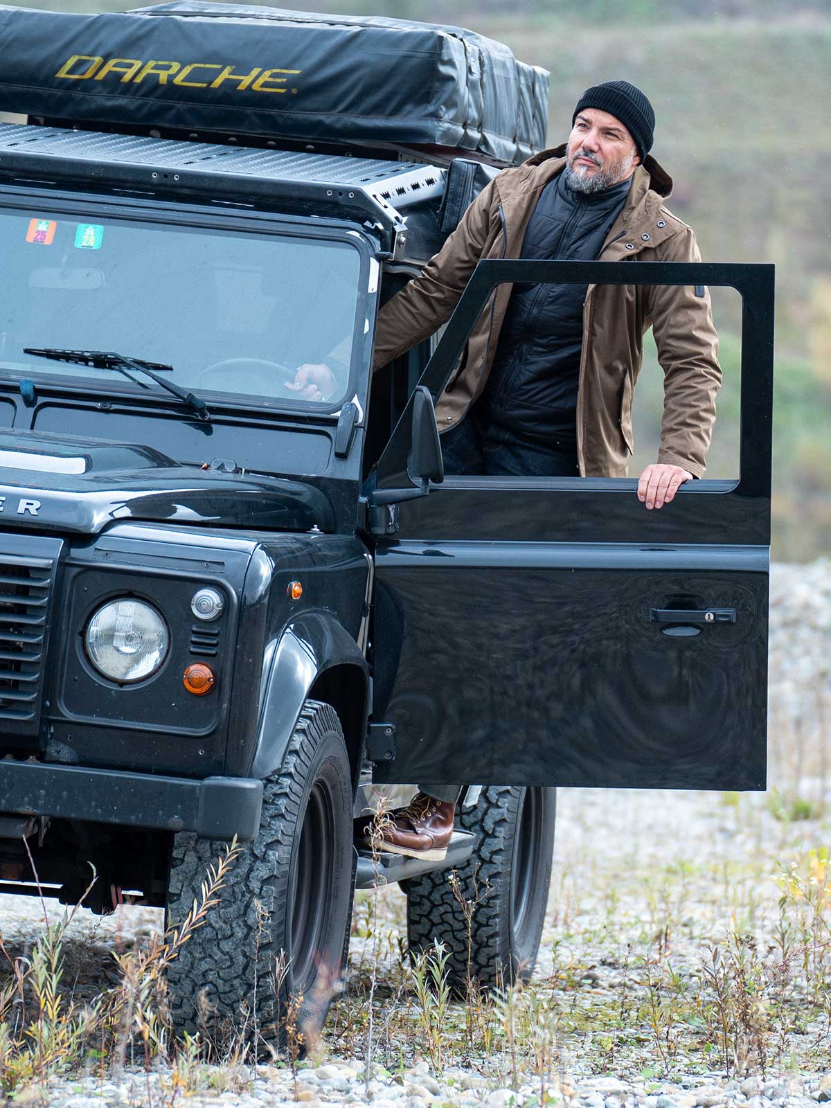 On-body shot of CADUFF Japanese Double-Knit 2-in-1 Winter Parka — Beech; outdoor mountain setting, model entering a black Land Rover Defender; premium winter jacket.