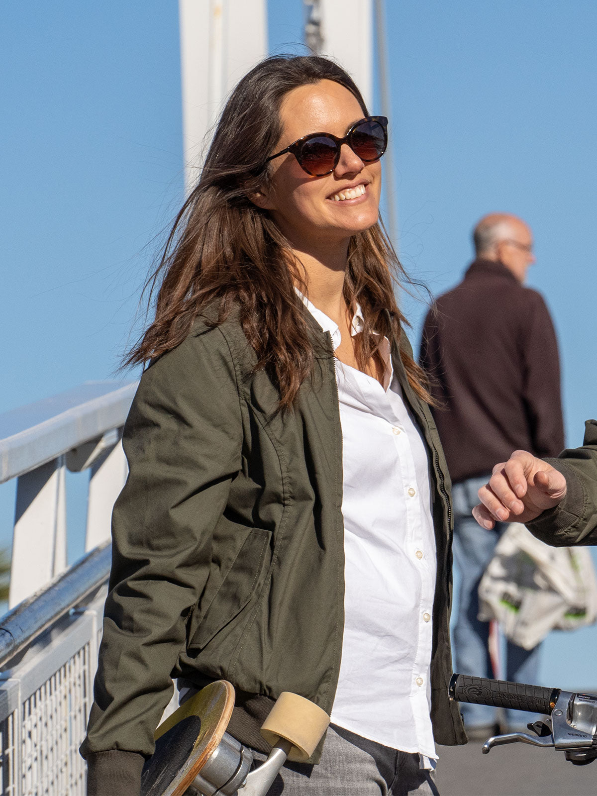 WOMAN MODEL WALKING WITH A SKATEBOARD IN THE CITY WEARING A GREEN URBAN MOBILITY BOMBER JACKET FOR LEGENDS BY CADUFF
