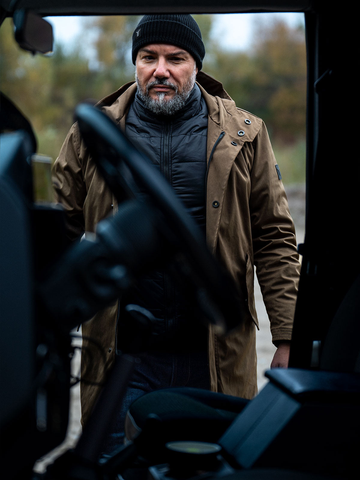 Interior view from a black Land Rover Defender: model approaches wearing CADUFF Japanese Double-Knit 2-in-1 Winter Parka — Beech, parka open with ECO DOWN liner visible; steering wheel in foreground.