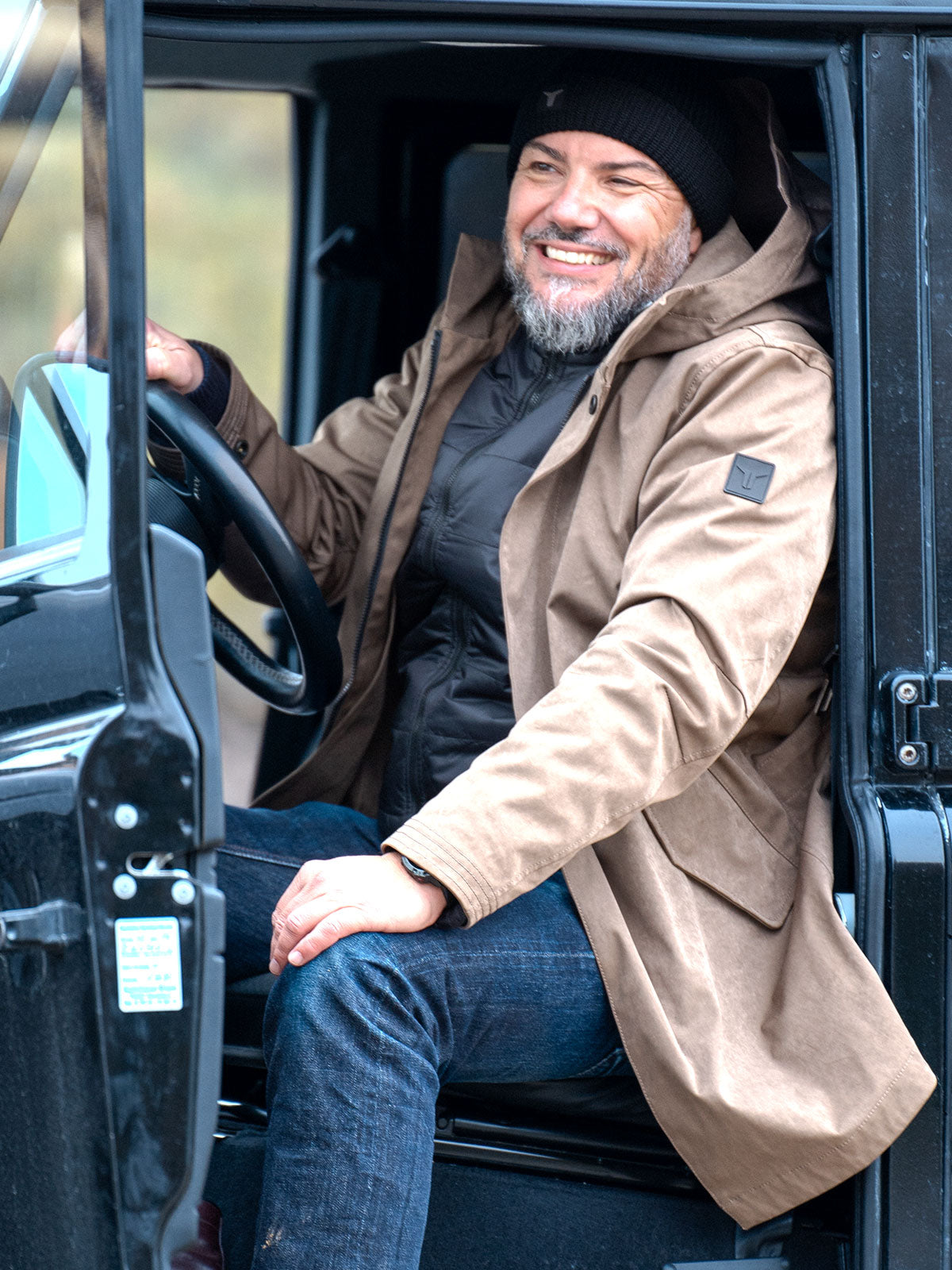 Interior shot: model seated in a black Land Rover Defender wearing CADUFF Japanese Double-Knit 2-in-1 Winter Parka — Brown; door open, ECO DOWN liner visible.