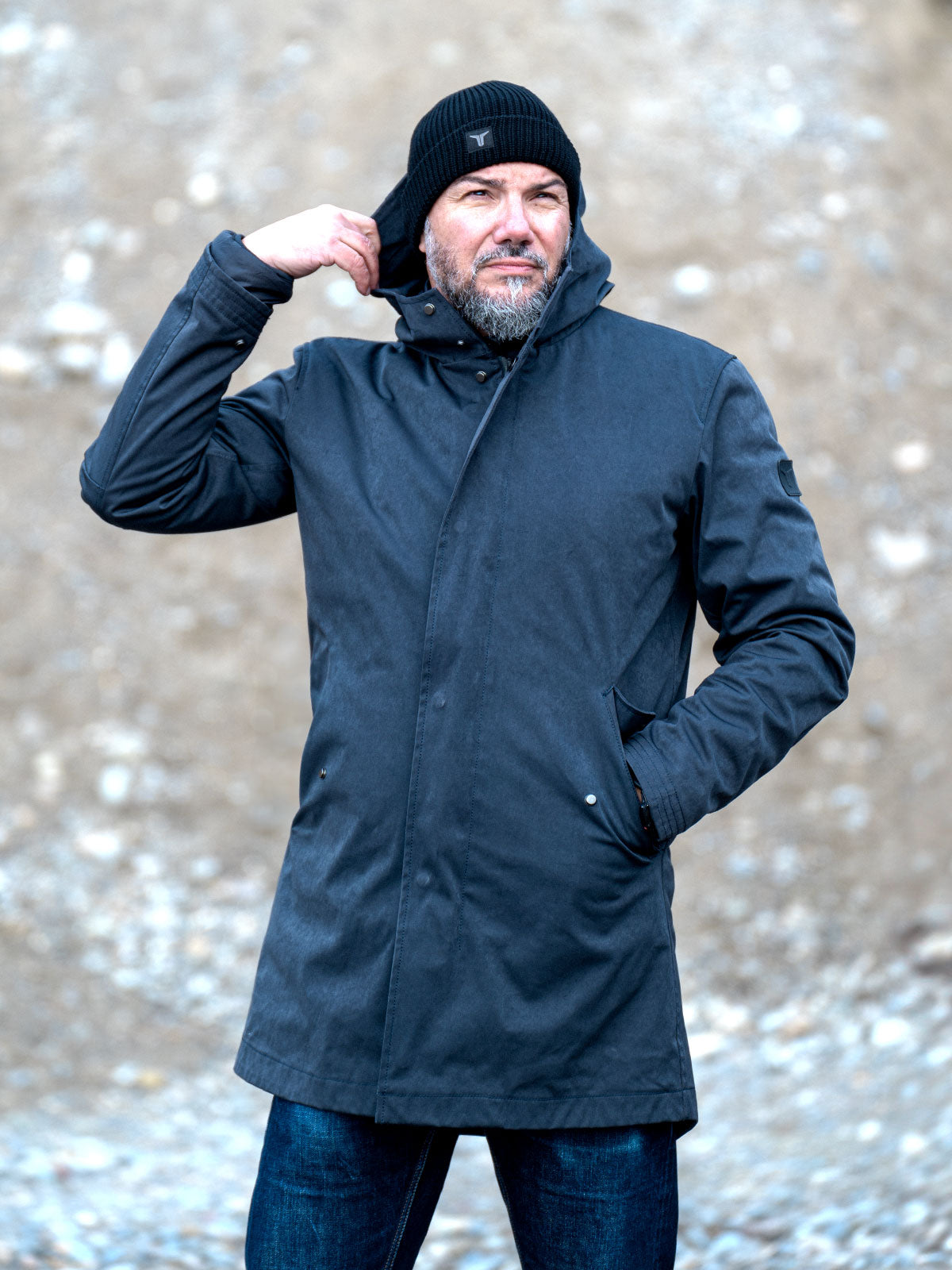 Model wearing a premium winter parka for men from for-legends.com by Caduff, crafted from exclusive high-density knit Japanese fabric, standing in a rocky winter landscape in the background a Land Rover Defender. / Modell trägt einen hochwertigen Winterparka für Herren von for-legends.com by Caduff aus einem exklusivem High-Density-Jersey aus Japan, stehend in einer felsigen Winterlandschaft im Hintergrund ein Landrover Defender.