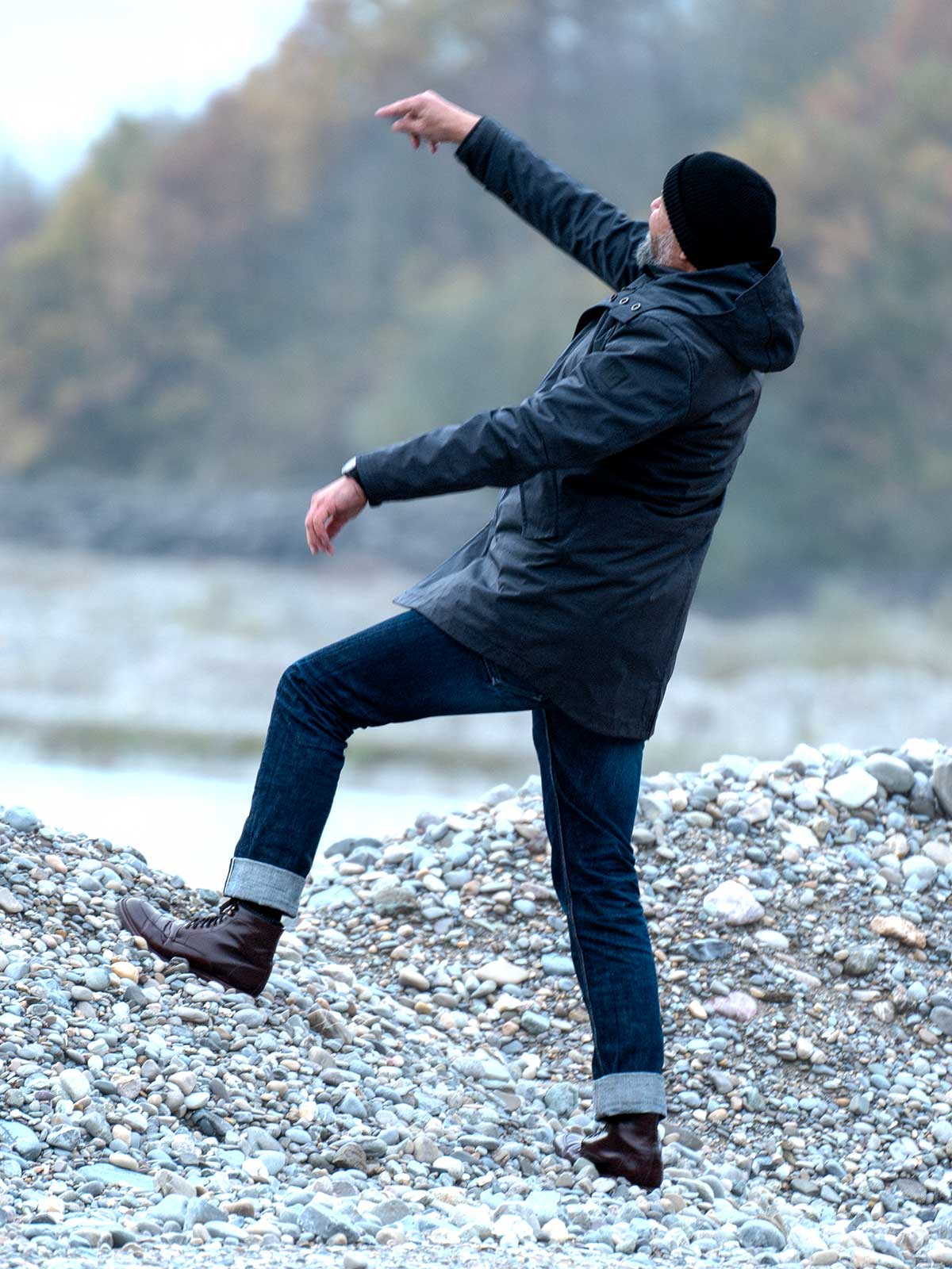 Model wearing a premium winter parka for men from for-legends.com by Caduff, crafted from exclusive high-density knit Japanese fabric, standing in a rocky winter landscape in the background a Land Rover Defender. / Modell trägt einen hochwertigen Winterparka für Herren von for-legends.com by Caduff aus einem exklusivem High-Density-Jersey aus Japan, stehend in einer felsigen Winterlandschaft im Hintergrund ein Landrover Defender.