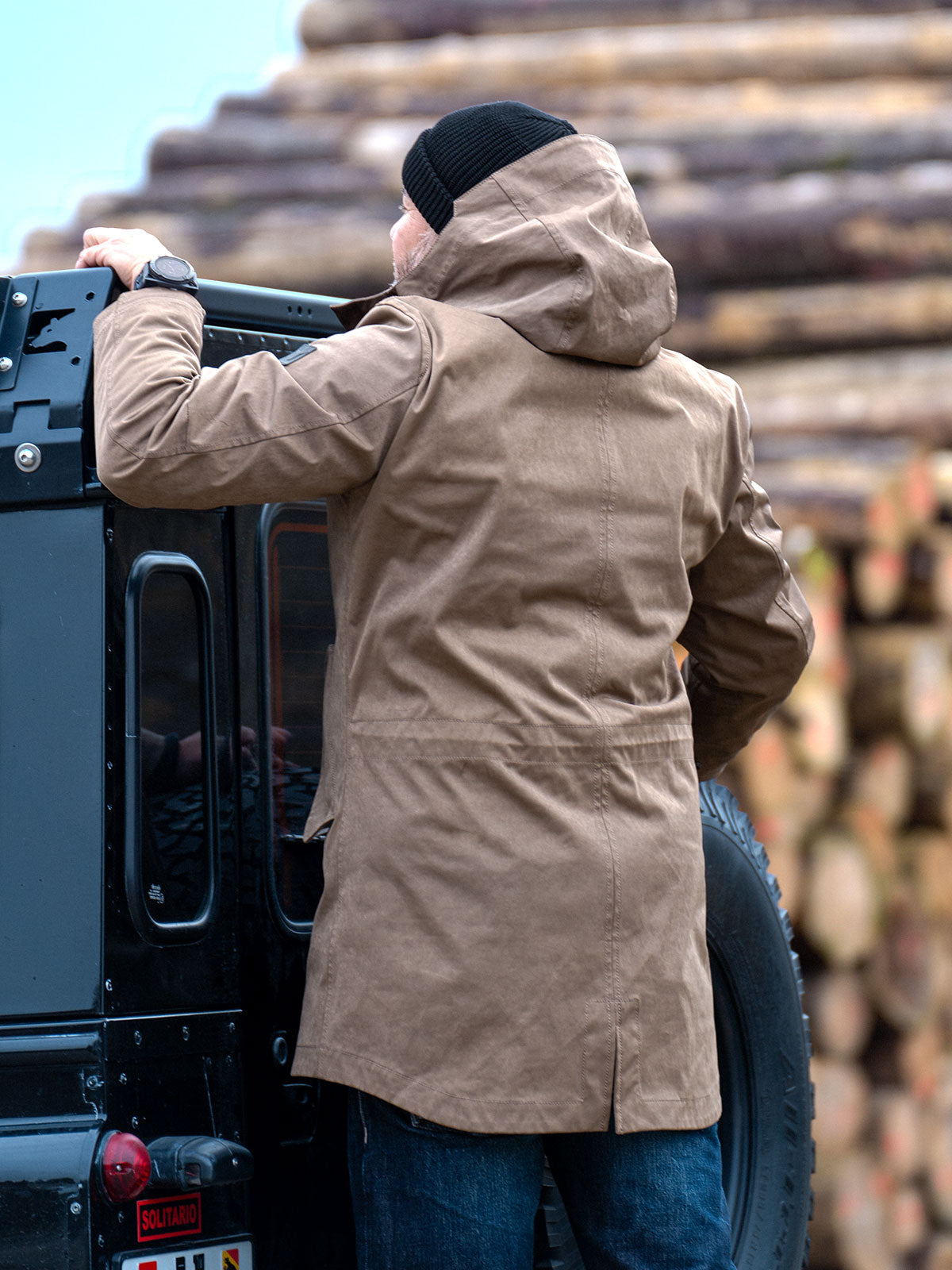 Model wearing a premium winter parka for men from for-legends.com by Caduff, crafted from exclusive high-density knit Japanese fabric, standing in a rocky winter landscape in the background a Land Rover Defender. / Modell trägt einen hochwertigen Winterparka für Herren von for-legends.com by Caduff aus einem exklusivem High-Density-Jersey aus Japan, stehend in einer felsigen Winterlandschaft im Hintergrund ein Landrover Defender.