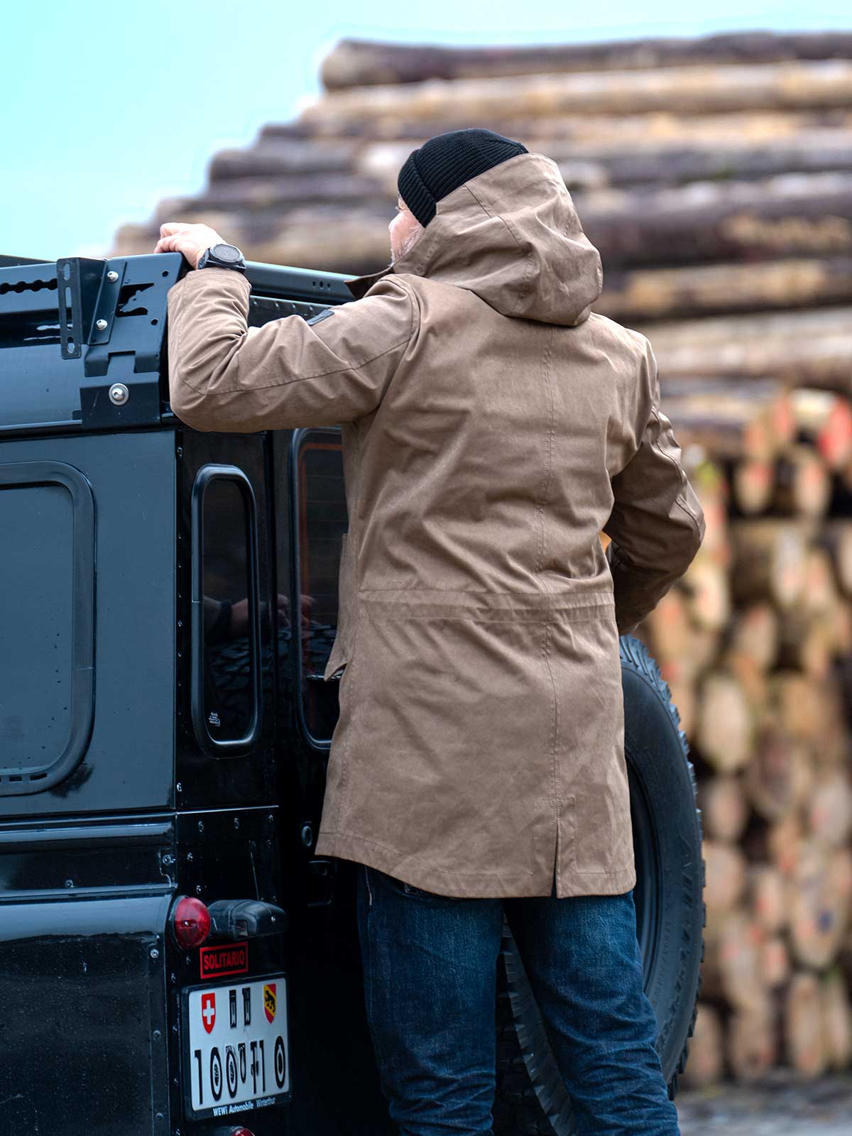 Model wearing a premium winter parka for men from for-legends.com by Caduff, crafted from exclusive high-density knit Japanese fabric, standing in a rocky winter landscape in the background a Land Rover Defender. / Modell trägt einen hochwertigen Winterparka für Herren von for-legends.com by Caduff aus einem exklusivem High-Density-Jersey aus Japan, stehend in einer felsigen Winterlandschaft im Hintergrund ein Landrover Defender.