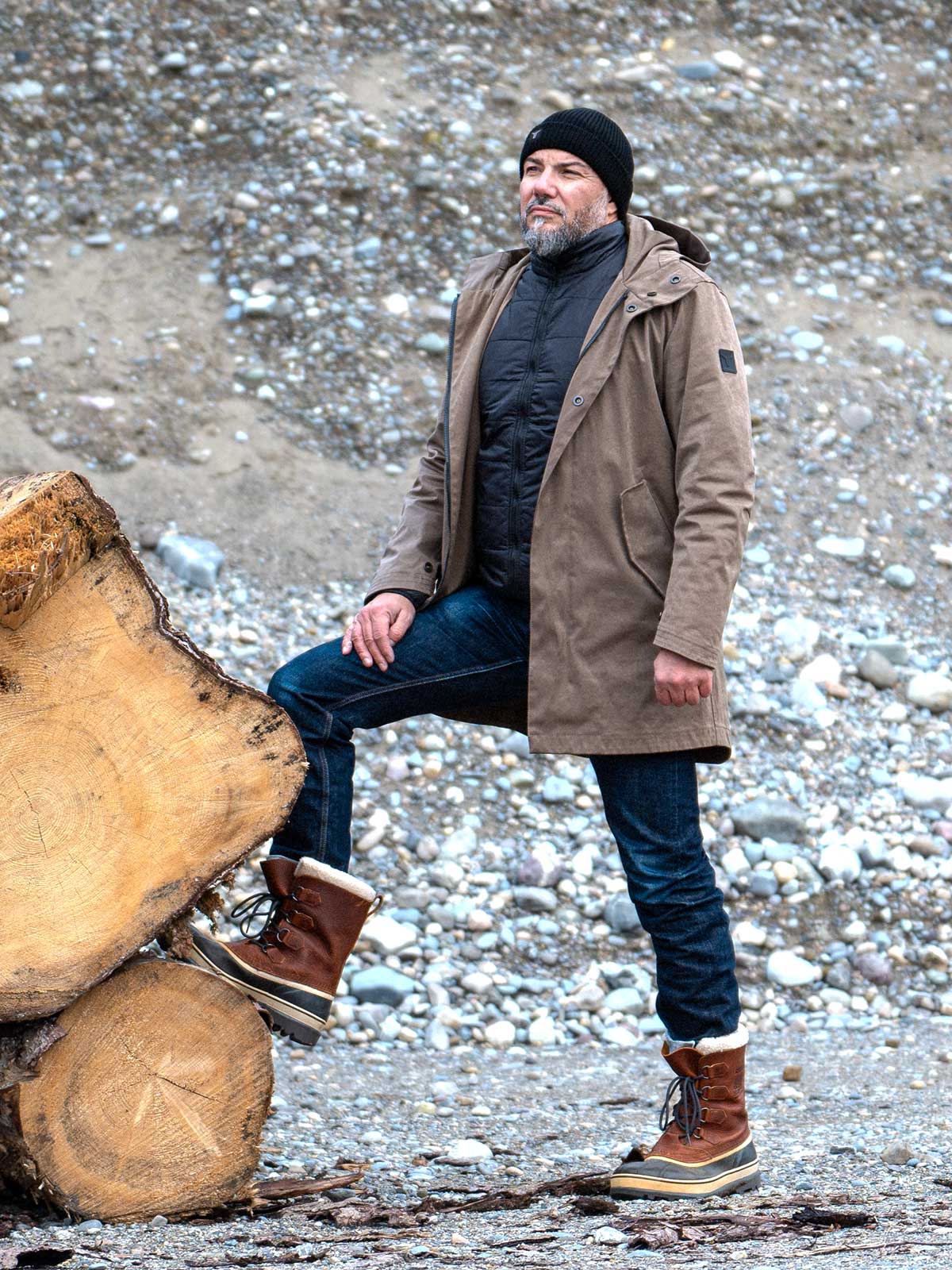 Model wearing a premium winter parka for men from for-legends.com by Caduff, crafted from exclusive high-density knit Japanese fabric, standing in a rocky winter landscape in the background a Land Rover Defender. / Modell trägt einen hochwertigen Winterparka für Herren von for-legends.com by Caduff aus einem exklusivem High-Density-Jersey aus Japan, stehend in einer felsigen Winterlandschaft im Hintergrund ein Landrover Defender.