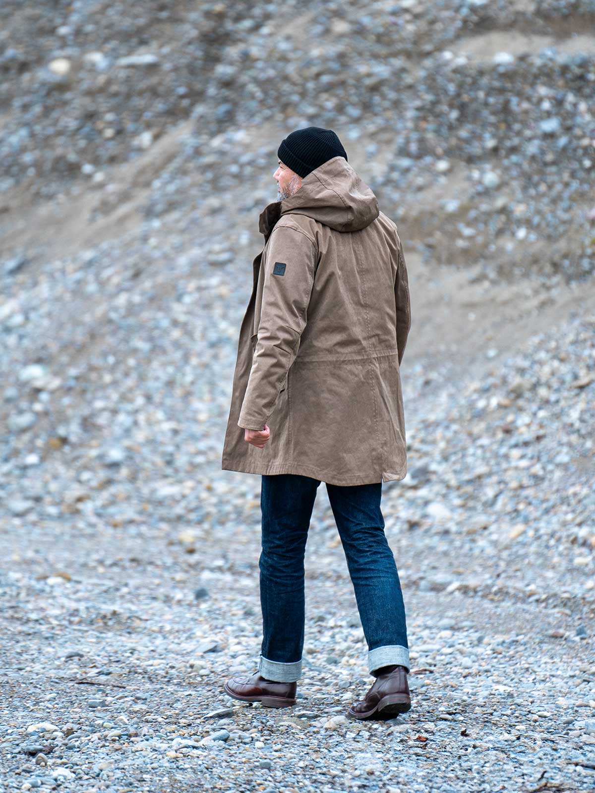 Model wearing a premium winter parka for men from for-legends.com by Caduff, crafted from exclusive high-density knit Japanese fabric, standing in a rocky winter landscape in the background a Land Rover Defender. / Modell trägt einen hochwertigen Winterparka für Herren von for-legends.com by Caduff aus einem exklusivem High-Density-Jersey aus Japan, stehend in einer felsigen Winterlandschaft im Hintergrund ein Landrover Defender.