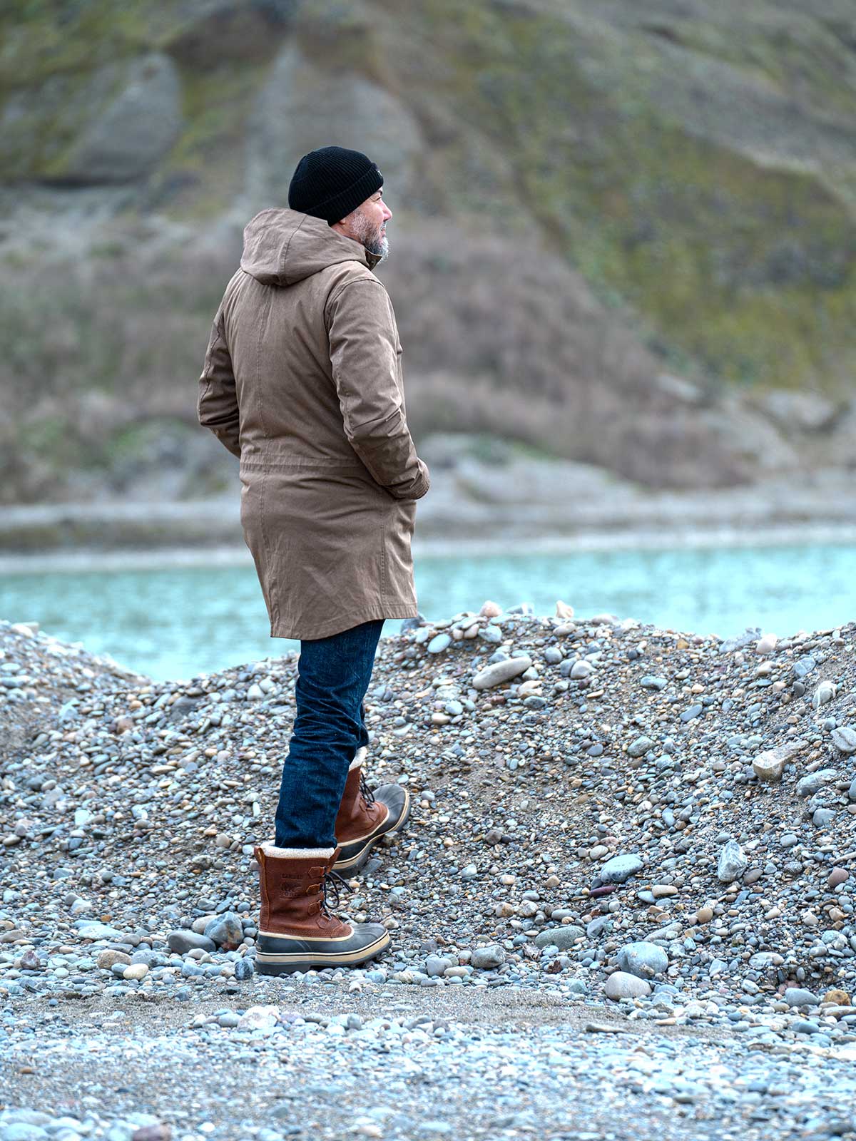 Model wearing a premium winter parka for men from for-legends.com by Caduff, crafted from exclusive high-density knit Japanese fabric, standing in a rocky winter landscape in the background a Land Rover Defender. / Modell trägt einen hochwertigen Winterparka für Herren von for-legends.com by Caduff aus einem exklusivem High-Density-Jersey aus Japan, stehend in einer felsigen Winterlandschaft im Hintergrund ein Landrover Defender.