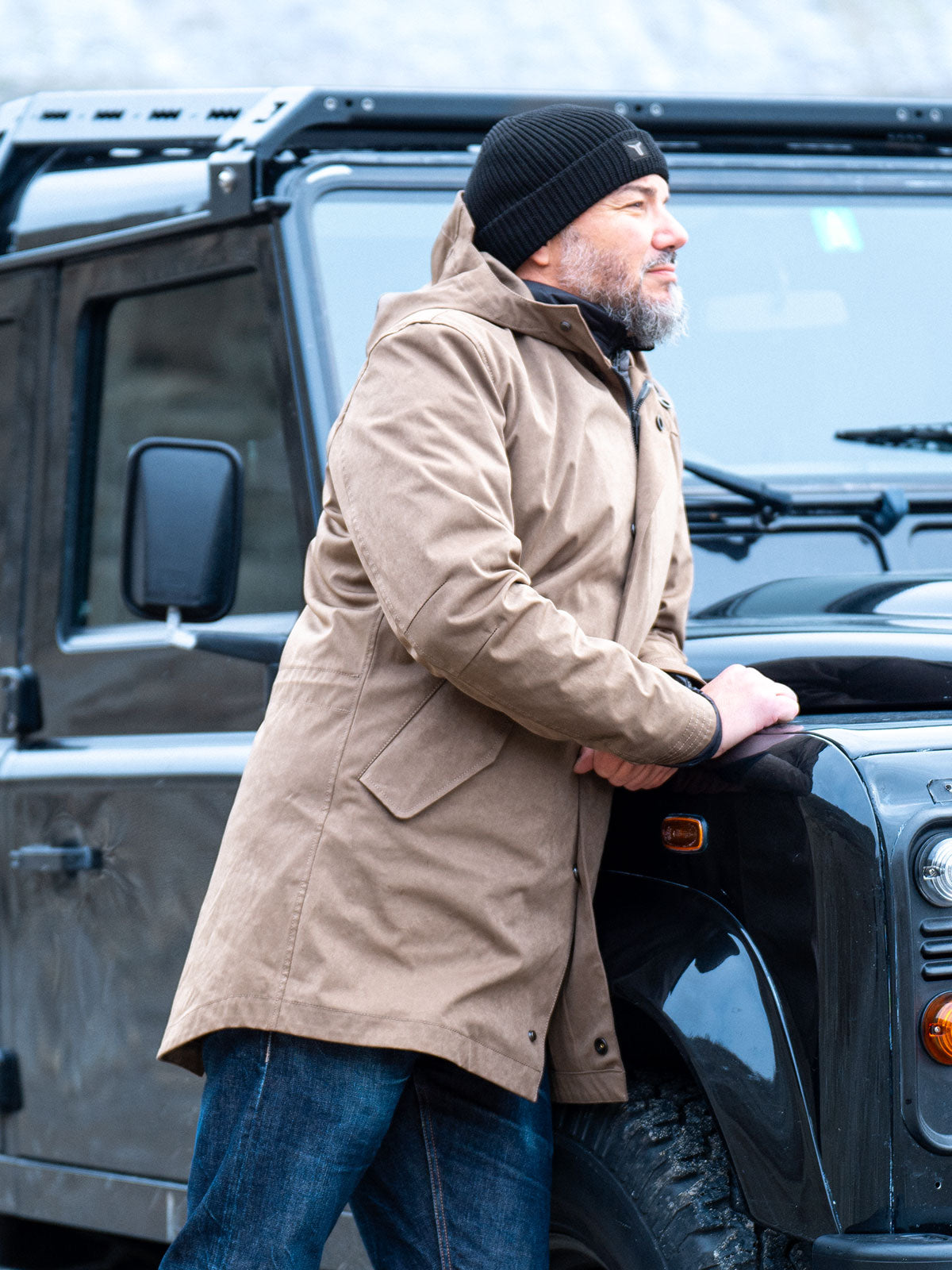 Model wearing a premium winter parka for men from for-legends.com by Caduff, crafted from exclusive high-density knit Japanese fabric, standing in a rocky winter landscape in the background a Land Rover Defender. / Modell trägt einen hochwertigen Winterparka für Herren von for-legends.com by Caduff aus einem exklusivem High-Density-Jersey aus Japan, stehend in einer felsigen Winterlandschaft im Hintergrund ein Landrover Defender.