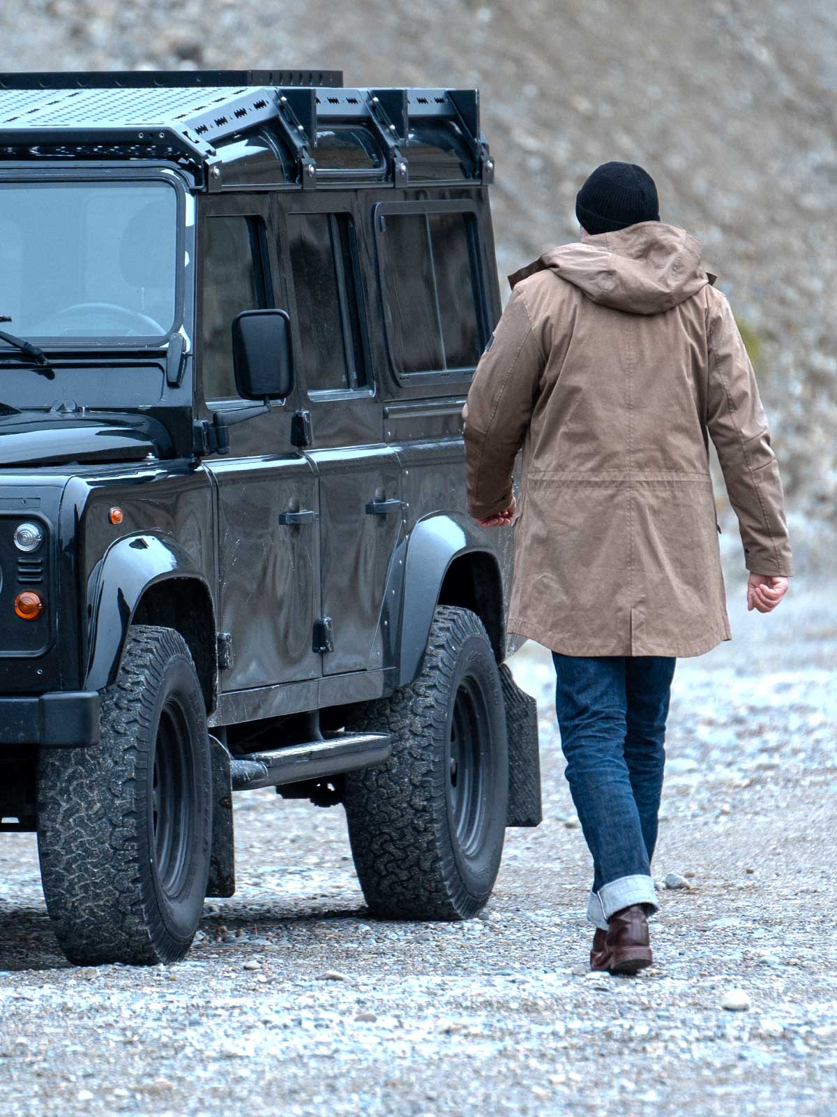 Model wearing a premium winter parka for men from for-legends.com by Caduff, crafted from exclusive high-density knit Japanese fabric, standing in a rocky winter landscape in the background a Land Rover Defender. / Modell trägt einen hochwertigen Winterparka für Herren von for-legends.com by Caduff aus einem exklusivem High-Density-Jersey aus Japan, stehend in einer felsigen Winterlandschaft im Hintergrund ein Landrover Defender.