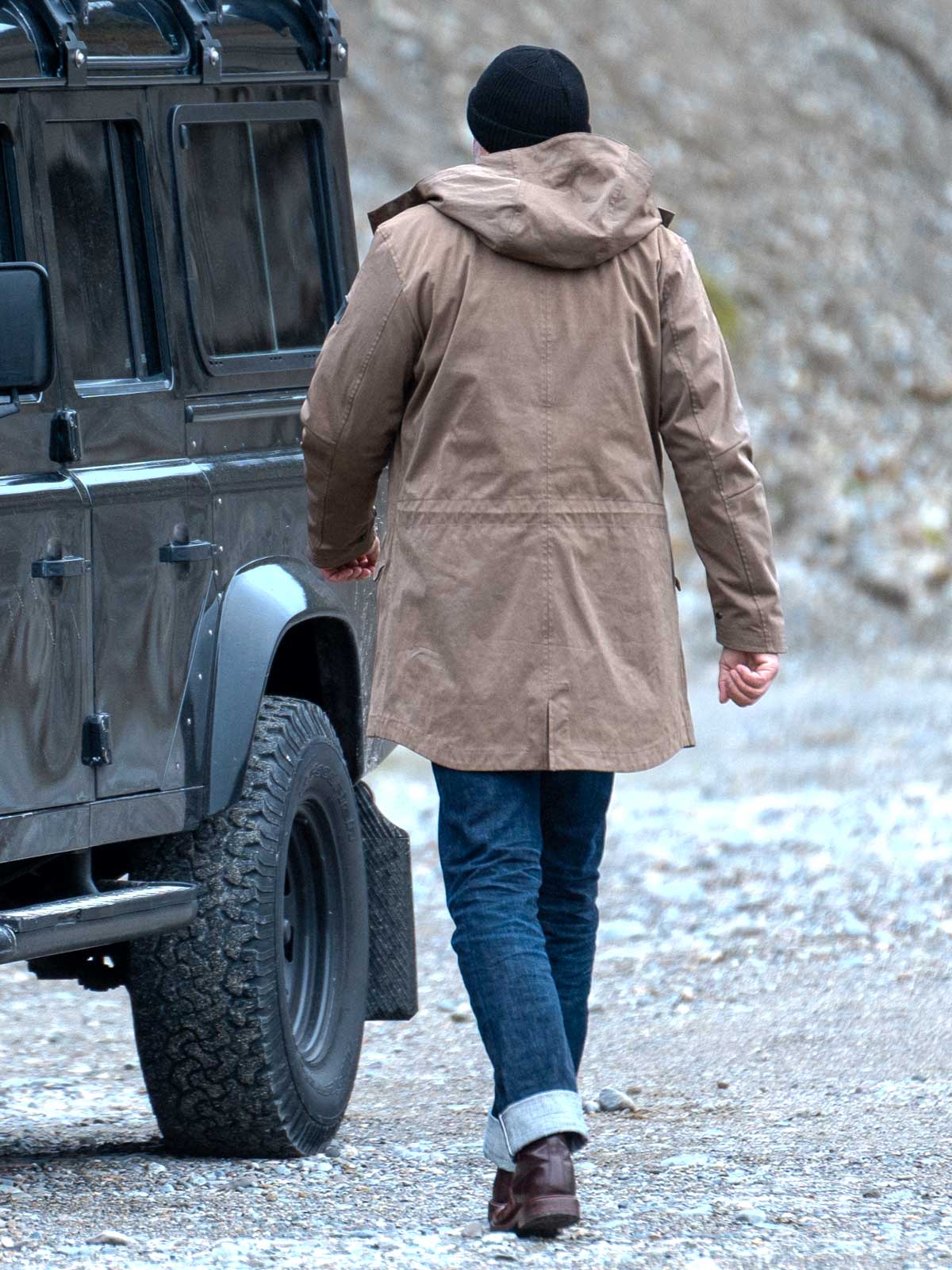 Model wearing a premium winter parka for men from for-legends.com by Caduff, crafted from exclusive high-density knit Japanese fabric, standing in a rocky winter landscape in the background a Land Rover Defender. / Modell trägt einen hochwertigen Winterparka für Herren von for-legends.com by Caduff aus einem exklusivem High-Density-Jersey aus Japan, stehend in einer felsigen Winterlandschaft im Hintergrund ein Landrover Defender.