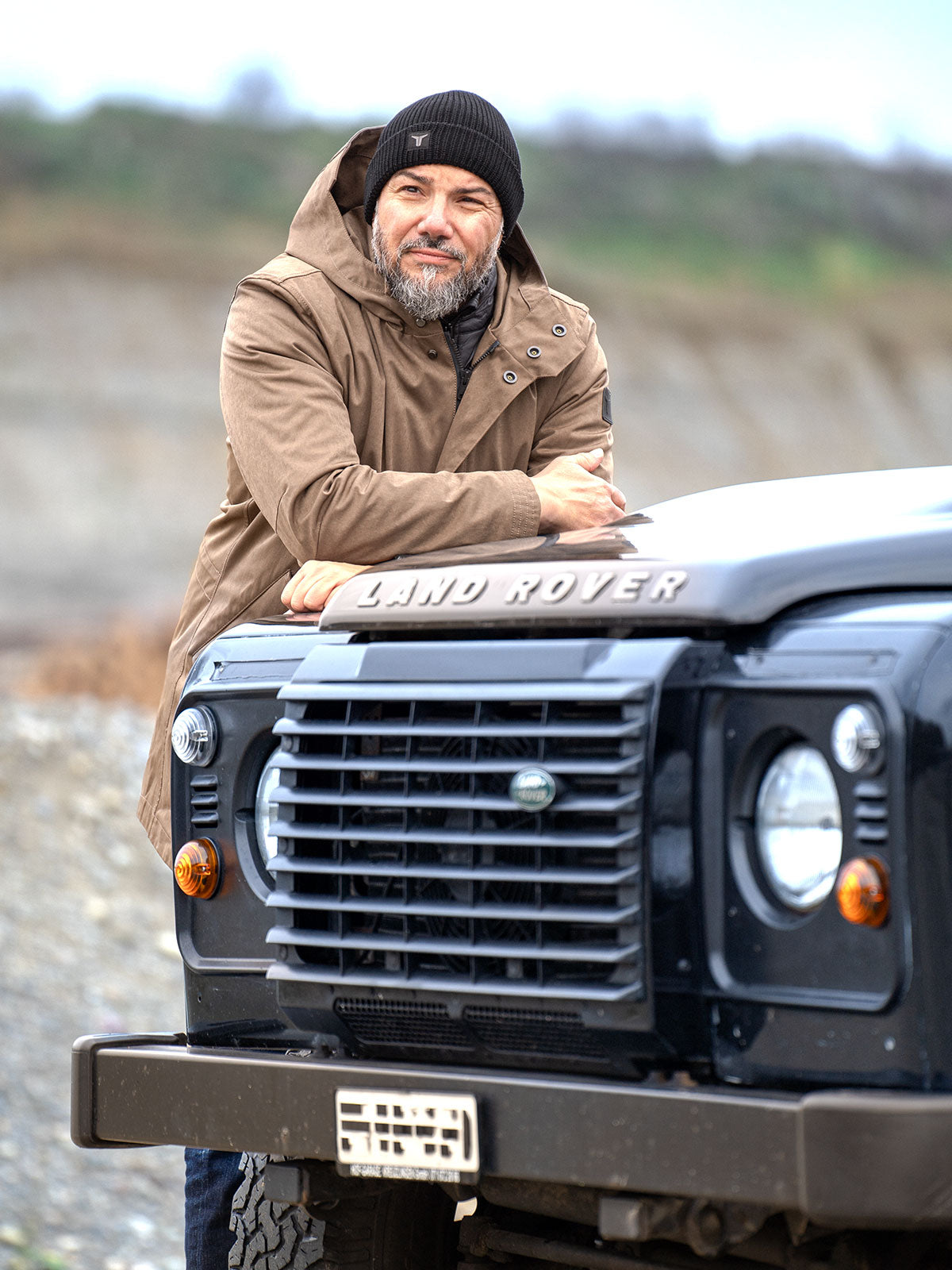 Model wearing a premium winter parka for men from for-legends.com by Caduff, crafted from exclusive high-density knit Japanese fabric, standing in a rocky winter landscape in the background a Land Rover Defender. / Modell trägt einen hochwertigen Winterparka für Herren von for-legends.com by Caduff aus einem exklusivem High-Density-Jersey aus Japan, stehend in einer felsigen Winterlandschaft im Hintergrund ein Landrover Defender.