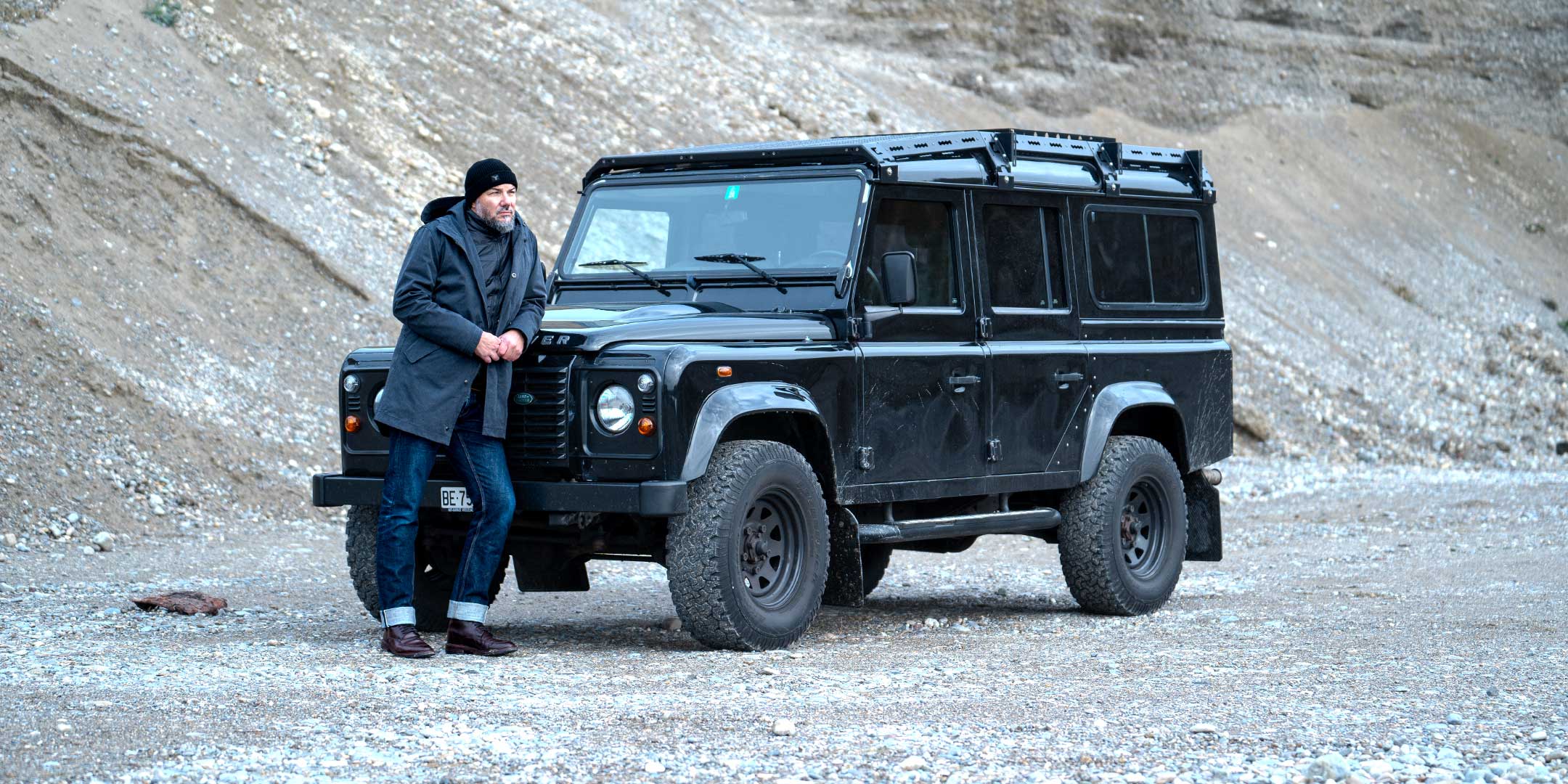 Model wearing a premium winter parka for men from for-legends.com by Caduff, crafted from exclusive high-density knit Japanese fabric, standing in a rocky winter landscape in the background a Land Rover Defender. / Modell trägt einen hochwertigen Winterparka für Herren von for-legends.com by Caduff aus einem exklusivem High-Density-Jersey aus Japan, stehend in einer felsigen Winterlandschaft im Hintergrund ein Landrover Defender.