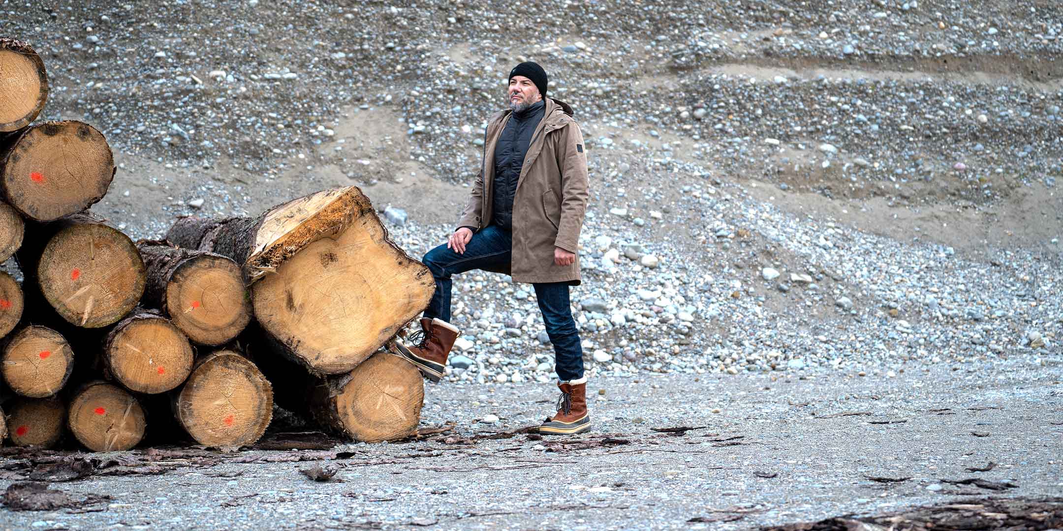 Model wearing a premium winter parka for men from for-legends.com by Caduff, crafted from exclusive high-density knit Japanese fabric, standing in a rocky winter landscape in the background a Land Rover Defender. / Modell trägt einen hochwertigen Winterparka für Herren von for-legends.com by Caduff aus einem exklusivem High-Density-Jersey aus Japan, stehend in einer felsigen Winterlandschaft im Hintergrund ein Landrover Defender.