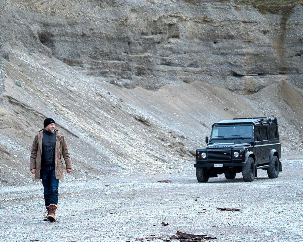 Brown CADUFF Japanese Double-Knit 2-in-1 Winter Parka, worn open with ECO DOWN liner visible; Land Rover Defender 110 on the right, mountains in the background.