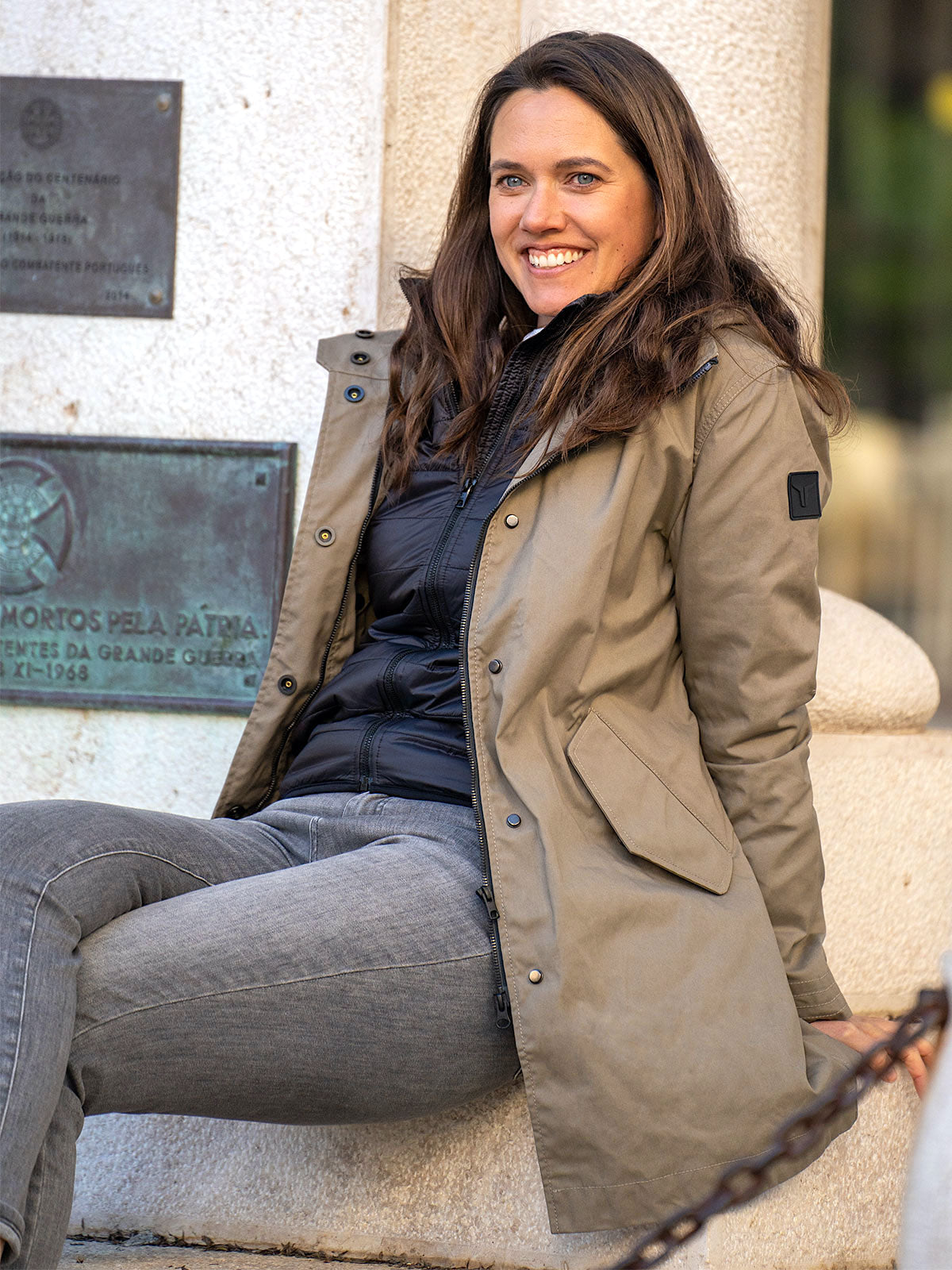 WOMAN MODEL SITTING  IN THE CITY IN FRONT OF A STATUE WEARING AN URBAN MOBILITY PARKA WITH AN ABRASSION RESISTANCE UP TO A SPEED OF 45 KM/H